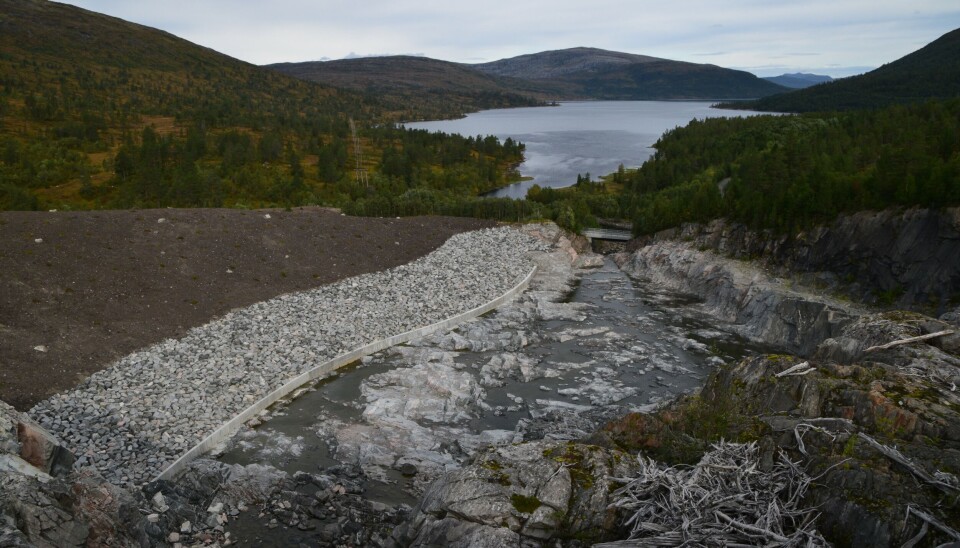 Nedstrøms Gråsjødammen er det lagt opp stein, som skal forhindre at jord vaskes bort når overløpet er på sitt kraftigste. Et fjellandskap som er endret etter anleggsarbeid. en bekk, og et fjellvann i bakgrunnen. Sommer og blå himmel.