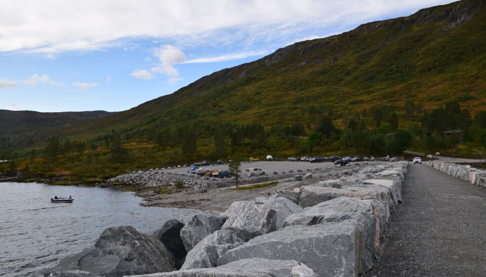 Fin ny parkeringsplass og båtutslipp ved Gråhaugen. Toppen av en stor demning på fjellet, en parkeringsplass i bakgrunnen, og en båt på vannet. Sommer og blå himmel.