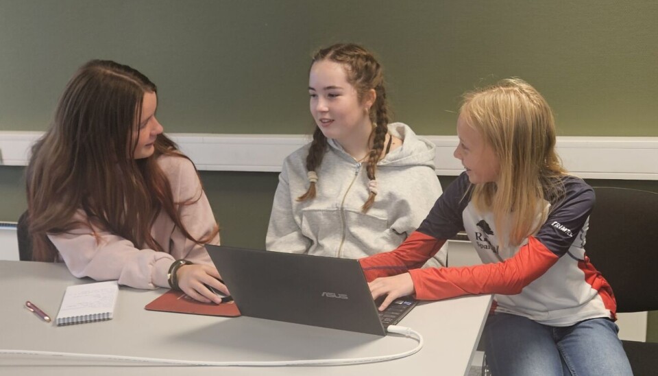 Her sitter vi og jobber med egen artikkel. Tre jenter som sitter på et rom og jobber med en artikkel på pc. De sitter ved et bord og en grønn vegg bak seg. En jente med lilla genser og mørkt hår, og en med grå genser og mørkt hår. Den siste jenta har en rød, blå og hvit genser med lyst hår.