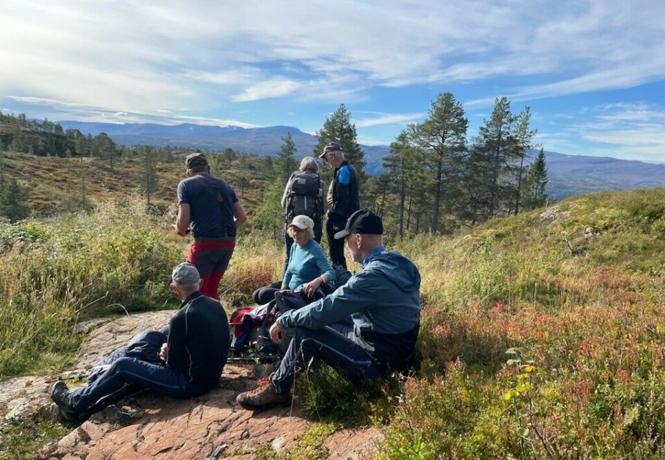 En liten pust i bakken. Seks friluftskledde personer på fjellet, tre av dem har satt seg ned for å hvile på et berg. Høst, sol og blå himmel.