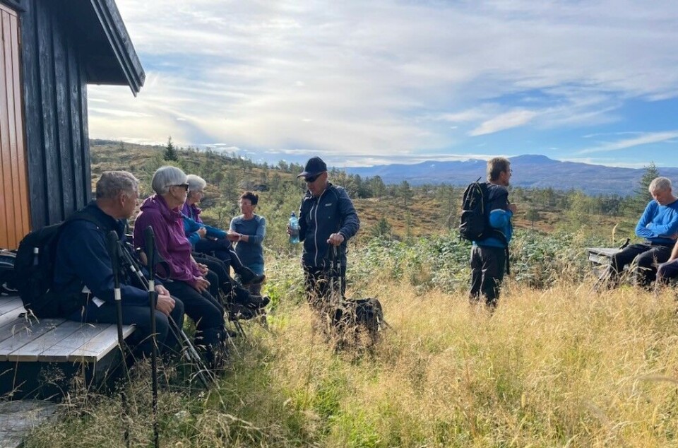 En liten pause før vi fortsetter mot toppen. Ei gruppe med friluftskledde folk som har samlet seg foran ei hytte i marka. Høst, sol og blå himmel.