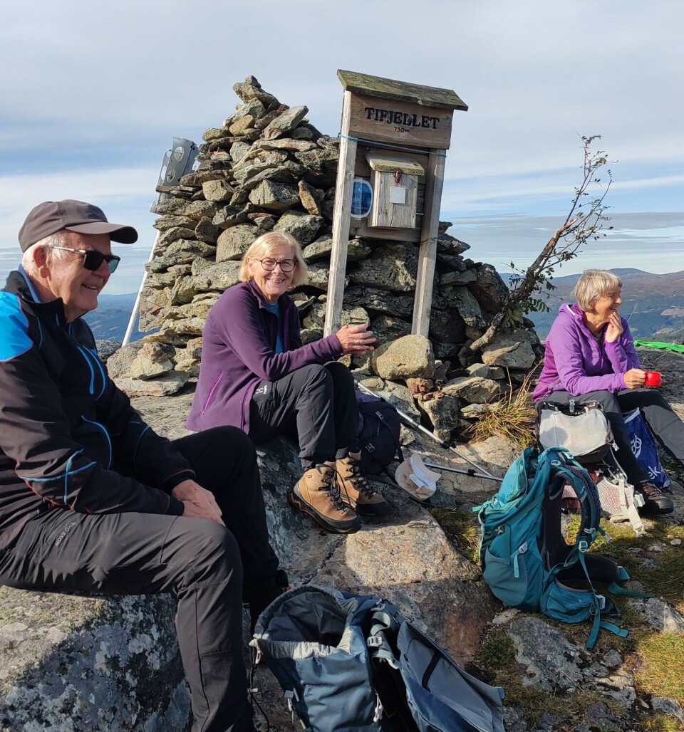 God stemning. Tre blide friluftskledde personer, en mann og to damer, som sitter og hviler ved en fjellvarde. Høst, sol og blå himmel.
