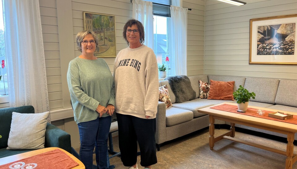 Kirsti Barbo Landsem og Anni Karlstrøm ønsker ALLE velkommen til Nonstreff på torsdager. To smilende damer som står foran en hjørnesofa inne i ei stue.