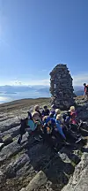 En gruppe barn sitter tetts ammen ved en varde på en fjelltopp. Blå himmel og litt skyer i bakgrunnen.