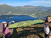 To unge jenter står på fjellet og peker ned på fjorden og gårer i dalen. Fjell og lett skyet himmel i bakgrunnen.