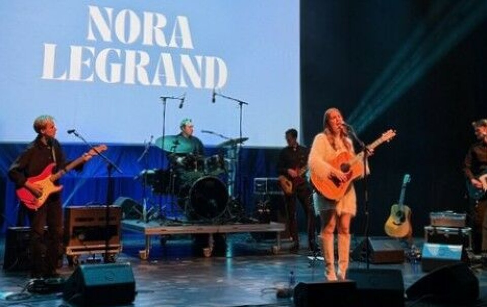 Nora Legrand. En kvinnelig sangartist med gitar på en scene. Bak henne et band.