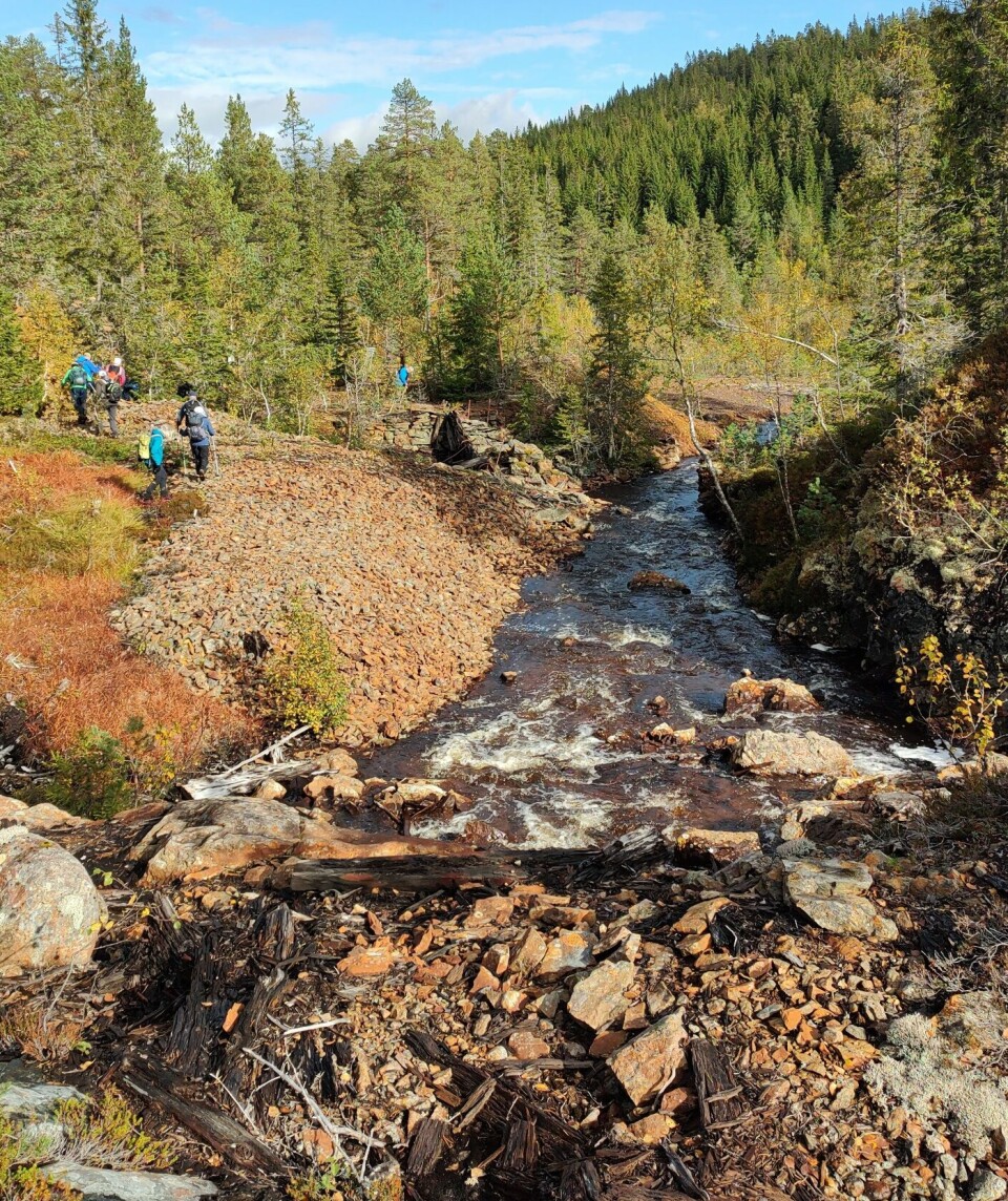 Tirsdagstur til Dragsetgruva En bekk som renner forbi slagghauger ett fortidens gruvedrift, og noen folk skimtes på avstand på sida av bekken. Høstvær og sol.