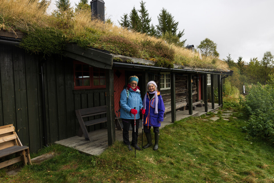 Idyllisk seter som har vorte koseleg turisthytte. To turkledde damer som står foran et gammelt seterhus med torvtak. Overskyet høstvær.