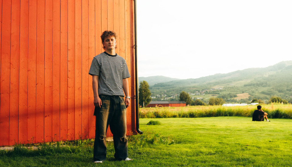 Jon Ranes til Surnadal Kulturhus. Bilet viser artisten Jon Ranes stående på en plen, foran en rød panelvegg, med utsikt mot åkrer og fjell i horisonten.