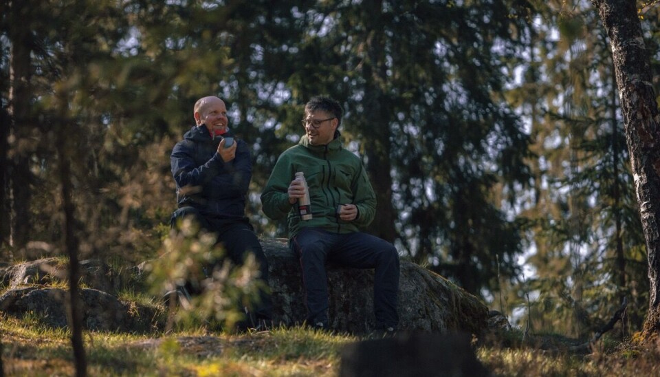 “Gjør noe fint for andre-dagen” 2. oktober Bildet viser to menn sittende på en stor stein i skogen, og det er mye skog, lyng og trær rundt. De sitter mede hver sin kaffekopp og termos, snakker sammen og ler.