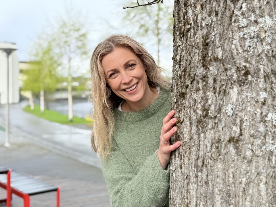 Mari Jørgensen Ryen, ABC-koordinator for Trøndelag Fylkeskommune Bildet viser en smilende kvinne med blondt hår og grønn genser, som titter ut fra bak et tre. I bakgrunnen er det fortau, trær, en benk og en lyktestolpe.