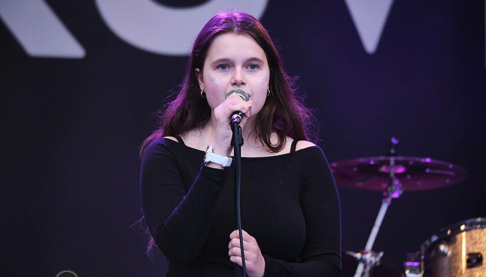Hedda Moen Gjerstad er en av debutantene for årets juleshow! Bildet viser en ung kvinne med mørkt løst hår stående på en scene. Hun synger inn i en mikrofon på et mikrofonstativ, og bakgrunnen og klærne hennes er mørke.