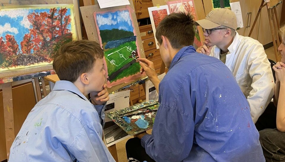 Kunstfagtime ved Rindal og Surnadal Kulturskole Bildet viser tre gutter med ryggen til kameraet. De sitter i store skjorter med malinmgsflekker på, og han i midten maler et maleri av en fotballbane. Guttene på høyre og venstre side tar seg til haken og følger med på prosessen. Bak maleriet er det mange staffeli, maleri og uferdige kunstprosjekter.