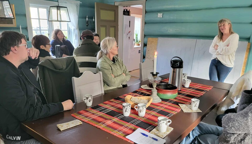 Gunn Elin på Holum gård. Ei dame som snakker til ei forsamling i ei gammel stue med tømmervegger. Tilhørerne sitter ved et gammelt langbord.