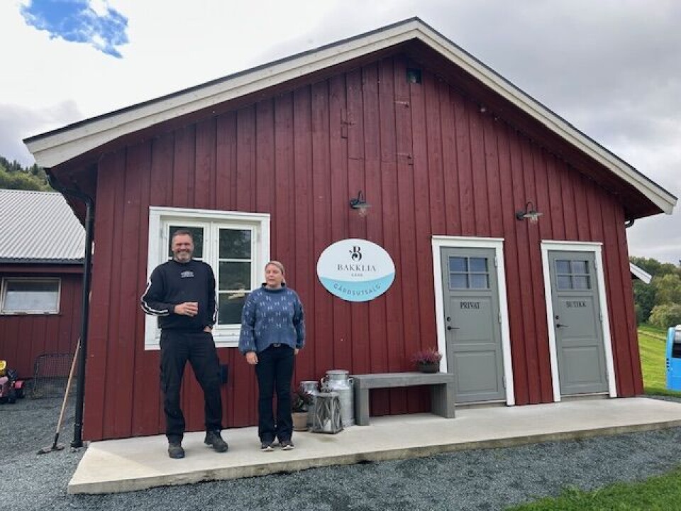Ola og Kristin på Bakklia gård. En mann og ei dame som står foran et rødmalt hus med et skilt på veggen.