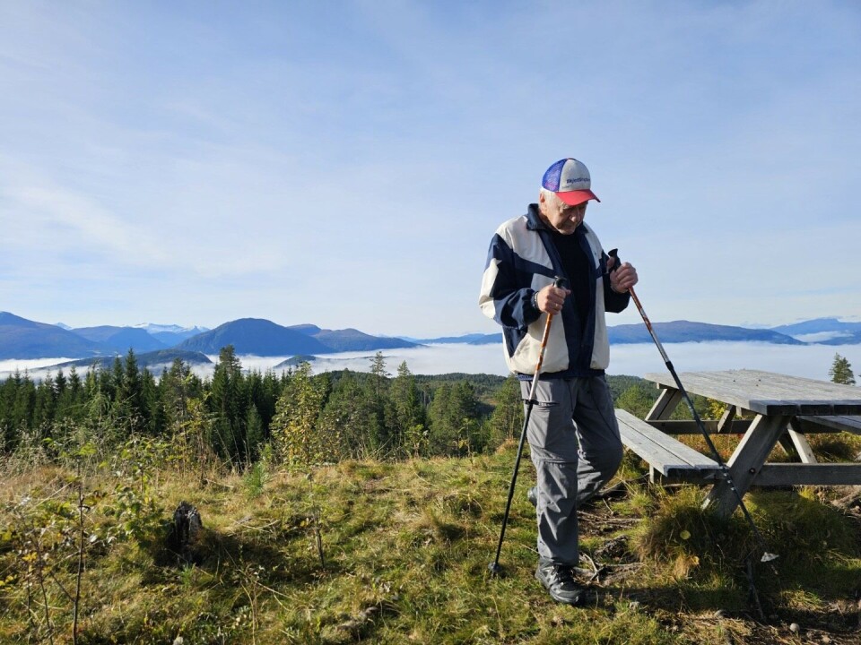 Sesongens siste stavgangtur En turkledd mann med grått hår og caps, og gåstaver, ved et rasteplassbord. Fjord og fjell i bakgrunnen, overskyet høstvær.