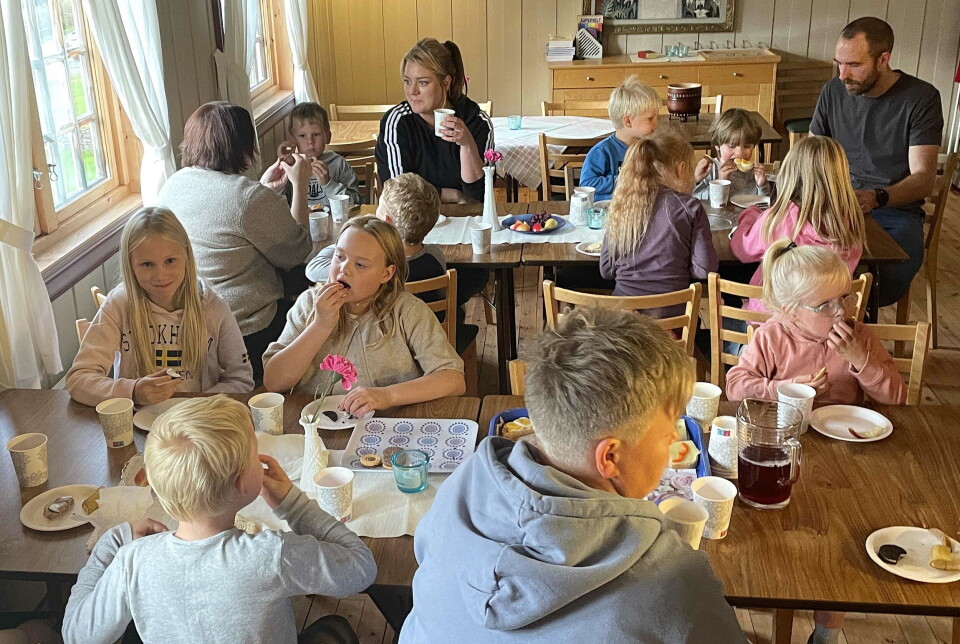 Alltid hyggeleg når Kari serverer i Kyrkjestua. Mange barn og noen voksne som sitter rundt to bord og spiser. Innendørs.