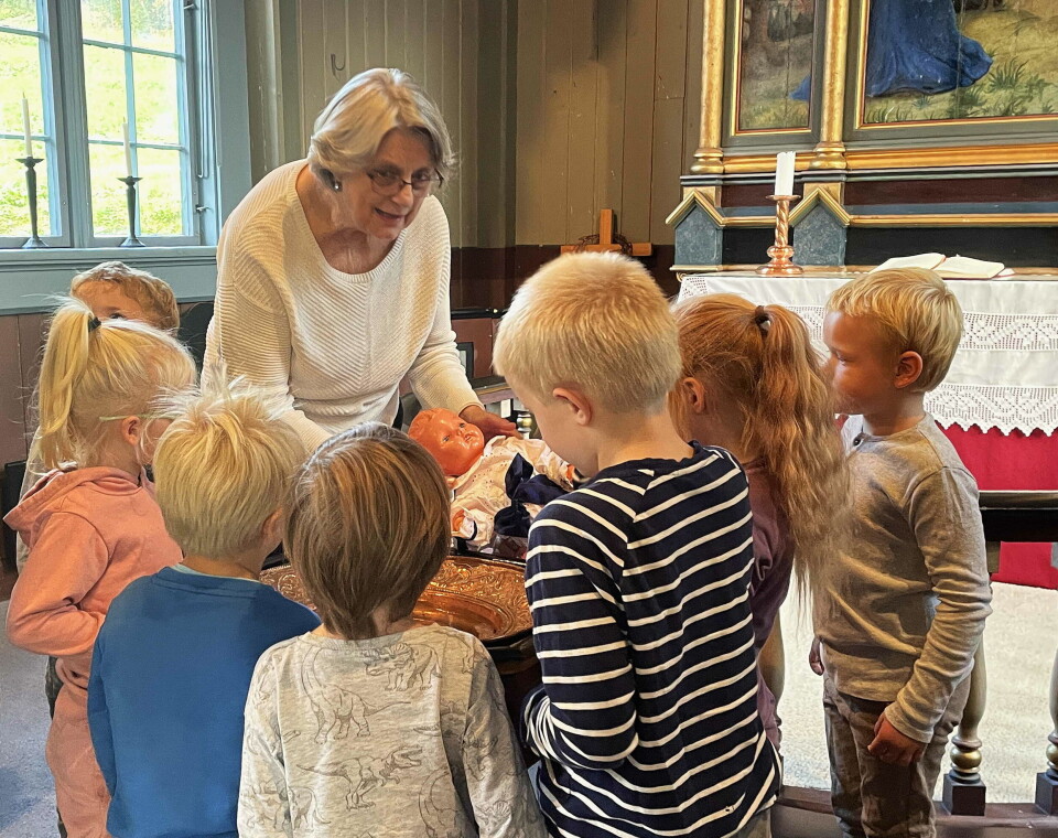 Leikedåp for lille «Sandra». Ivrige fadderar hjelper klokkar Randi – som leika prest. Ei dame med grått hår og briller som holder ei babydukke, og flere barn, rundt en døpefont i ei kirke.