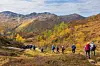 Ei gruppe med turgåere som går på rekke på en fjellsti. Høstfarger, sol og lettskyet himmel.