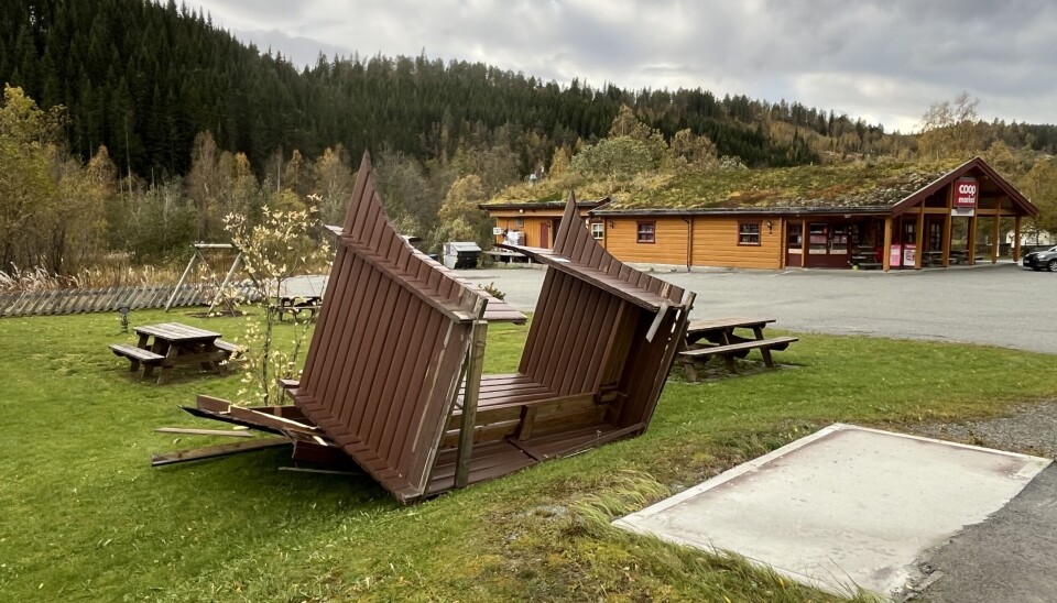 Vinden har veltet busskuret ved Nybø Bildet viser et busskur som står på hodet, med en dagligvarebutikk i bakgrunnen.