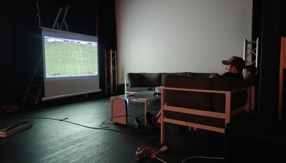 FIFA-turneringen foregikk oppå scenen på stort lerret. Bildet er av to menn sittende i en sofa, sett fra siden. De sitter oppå en scene og spiller fotballspillet FIFA på et stort lerret, ellers i rommet er det mørkt.