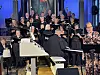 Et sangkor med blanding av menn og kvinner. Til høyre i bildet en dame som synger i en microfon. Foran i bilde et hvitt orgel og en mann som spiller piano ved siden av