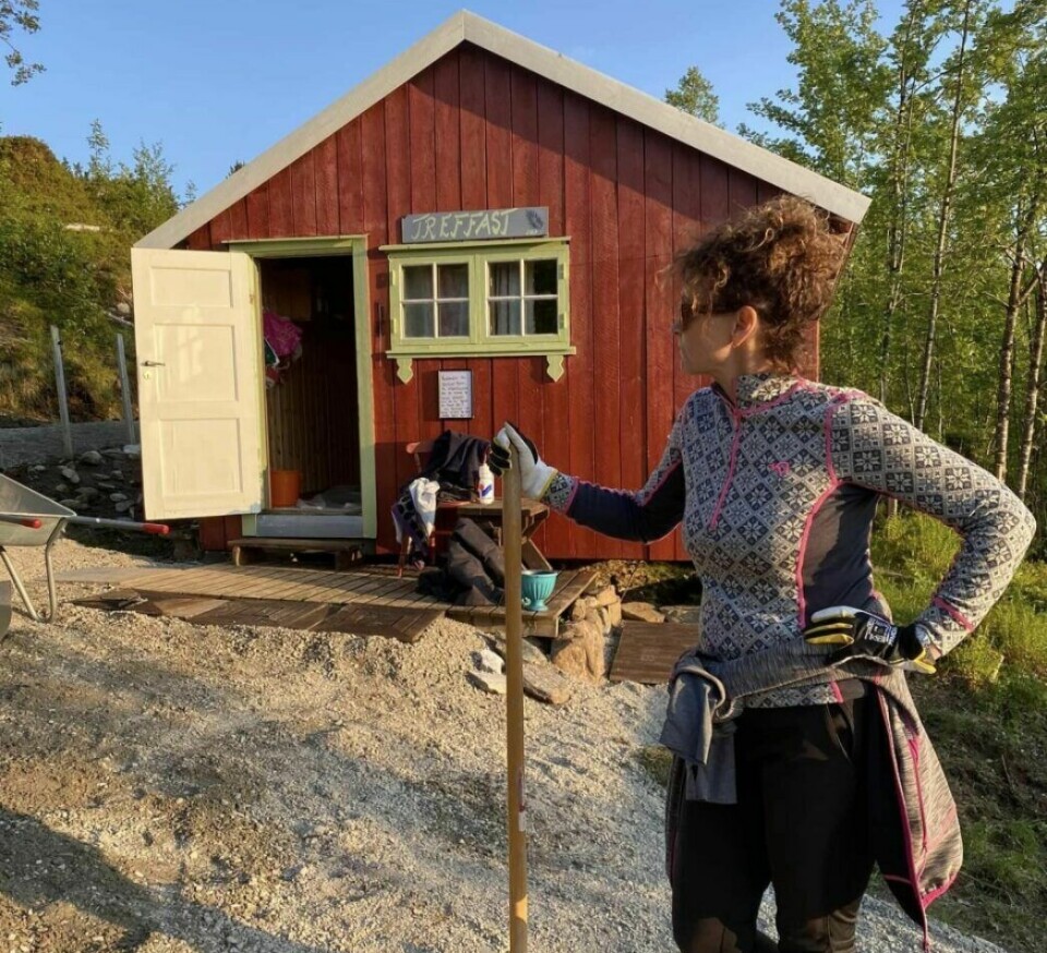 Treffasthytta Ei dame som står med ei krafse foran ei lita hytte, i solskinn.
