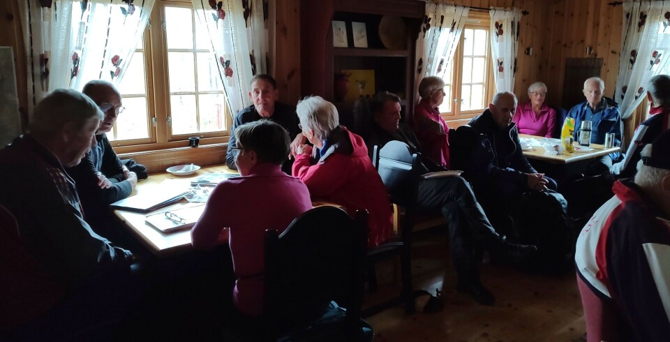 Sesongens siste tirsdagstur gikk til Gammelseterøyan Ei stor gruppe med turkledde folk som sitter trangt rundt to spisebord ved vinduer inne i ei hytte med trehvitt panel på veggene.