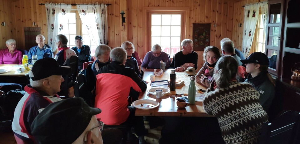 Sesongens siste tirsdagstur gikk til Gammelseterøyan Ei stor gruppe med turkledde folk som sitter trangt rundt to spisebord inne i ei hytte med trehvitt panel på veggene.