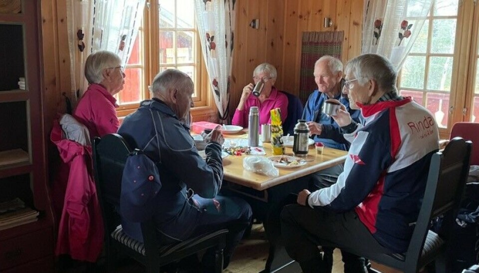 Sesongens siste tirsdagstur gikk til Gammelseterøyan Ei gruppe med fem turkledde personer som sitter rundt et spisebord ved viduet i ei hytte med trehvitt panel på veggene.