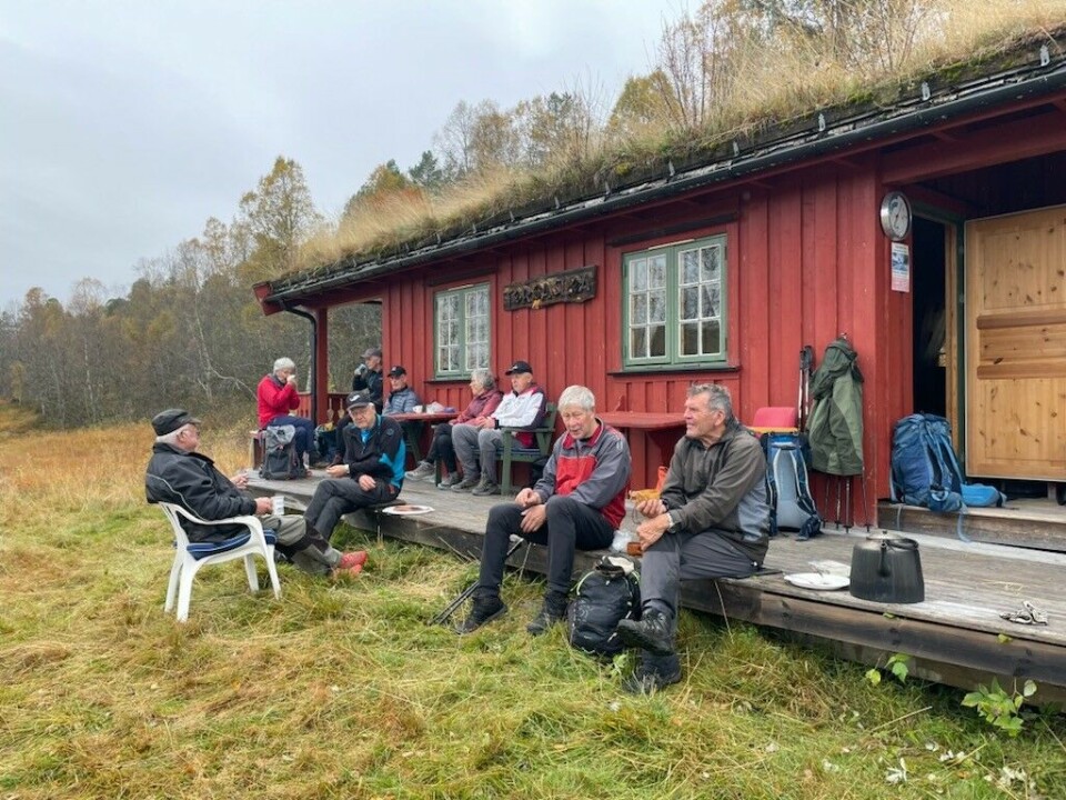 Sesongens siste tirsdagstur gikk til Gammelseterøyan Ei gruppe med turgåere som har tatt en pause ved ei rødmalt hytte med tovtak i marka. Overskya høstvær.