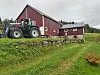 En mann som står i stigtrinnet på en traktor foran et fjøs. På tunet er det grønt gress, og himmelen er overskyet.
