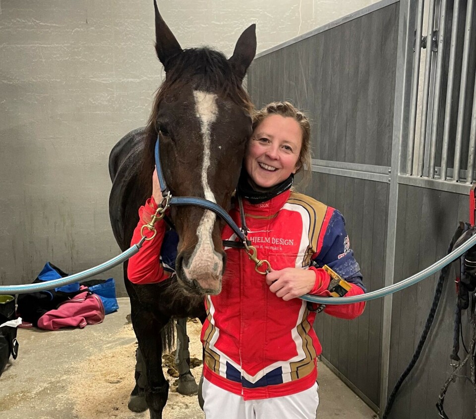 Scarselli med rytter Marit Valstad. Ei smilende dame med kuskedrakt, som står ved hodet til en travhest i en oppstallingsboks.