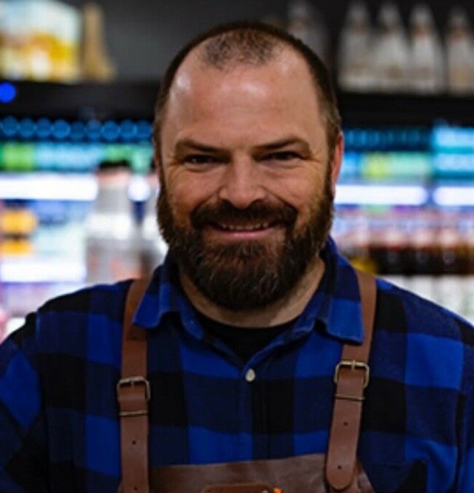 Tommy Holen Helland. Portrettbilde av en smilende mann med mørkt skjegg og veldig kort hår. Han har blå og svart rutete flanellskjorte på seg, og et forkle med lærremmer over skuldrene.