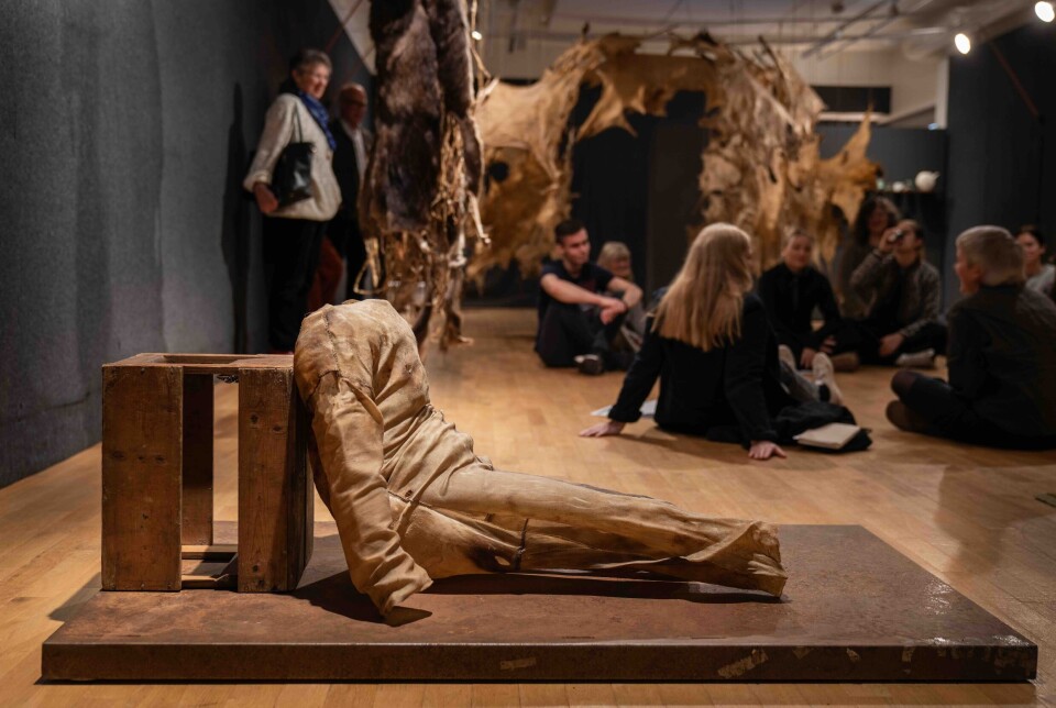 Frå omvising i utstillinga Doggerland ved Møre og Romsdal Kunstsenter. Utstillinga står til 1. november. En kunstutstilling. En figur som forestiller et menneske uten hode. Flere personer sitter i ring på gulvet i bakgrunnen.