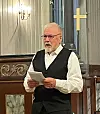 En mann med grått hår og skjegg og briller, ikledd hvit skjorte, svart bukse og svart vest, som står og leser opp noe fra et ark i ei kirke.
