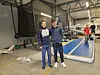 Bildet viser to unge menn stående i et stort lagerbygg. Bak dem ser vi en stor trampoline med nett, og en lang turnmatte på gulvet.