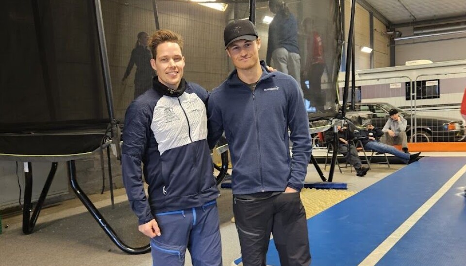 Ansvarlige for trampolinetrenerkurs Kjetil Øygard og Samuel Jøssang Bildet viser to unge menn stående i et stort lagerbygg. Bak dem ser vi en stor trampoline med nett, og en lang turnmatte på gulvet.