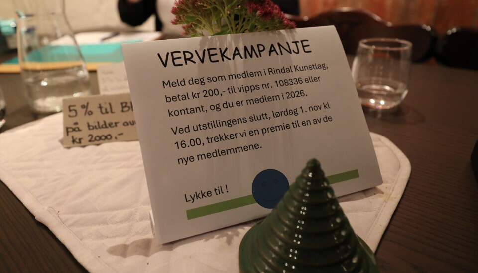 Kunstlaget sin vervekampanje. Melder du deg inn i løpet av utstillingen, kan du vinne en premie når utstillingen avsluttes. Bilde av plakat på bord med vervekampanje-tekst. LIte grønt keramikktre i forgrunnen.