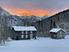To gamle bygninger, trær, fjell og solnedgang på himmelen. Skyer og sne.