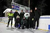 En dame og sju menn i varme klær foran vantet på en ishockeybane, sponsorskilt i bakgrunnen