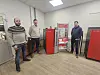 Tre personer står ved røde biofyringsanlegg i et utstillingsrom hos Biomentek AS.
