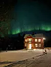Rødt hus i snølandskap under sterkt grønt nordlys på nattehimmel.