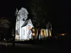 Hvit kirke opplyst om natten med lys i vinduene og mørke trær rundt.