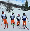 Fire barn med startnummer og skisportutstyr står i snødekt alpinbakke.