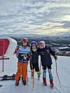 Tre barn i alpintutstyr står tett sammen i skibakken med fjell i bakgrunnen.