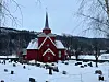 Rød trekirke med spiret midt i en snødekt kirkegård om vinteren.