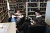 To personer sitter i svarte lenestoler og leser bøker mellom høye bokhyller på et bibliotek.