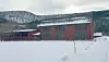 Stor rød bygning ved snødekt jorde med skogkledde åser i bakgrunnen.