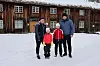 Familie i vinterklær står på snø foran gammel tømmerbygning